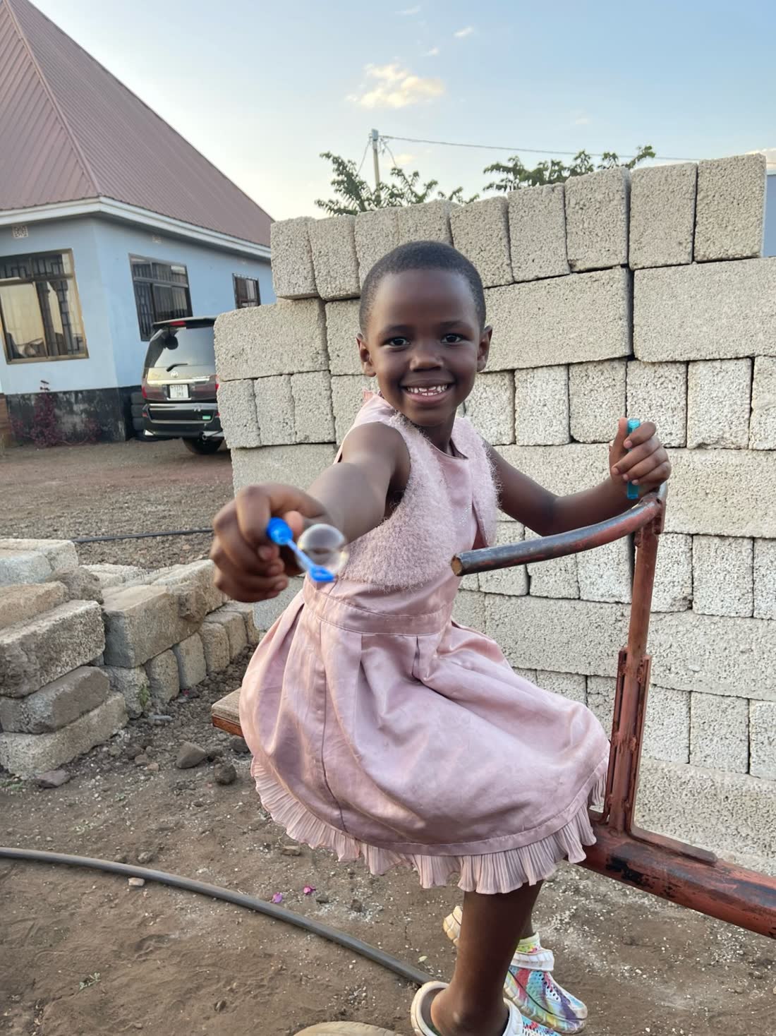 A girl in a pink dress at the home blowing a soap bubble in the yard at the end of the day.