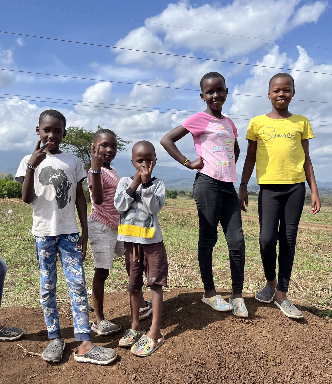 Children from Holley Children Orphanage standing together on red earth in an open field outside Arusha, under a wide sky with the hills behind them.