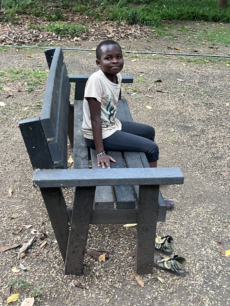 A boy sitting sideways on a park bench with his shoes kicked off beside it.