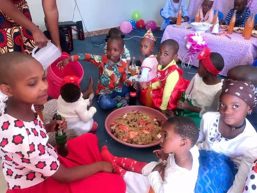 Children in party hats sitting in a circle on the floor around a platter of pilau, with balloons behind them.
