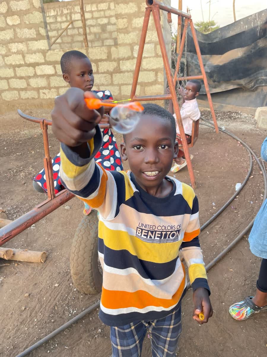 A boy holding out a bubble wand in the yard, with two children on the swings behind him.