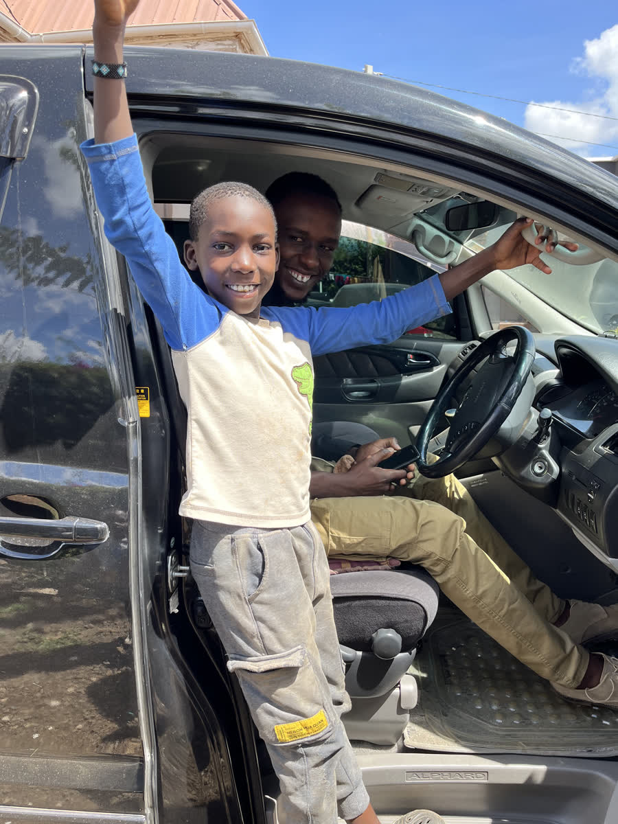 A boy standing in an open car door with both arms raised, laughing.