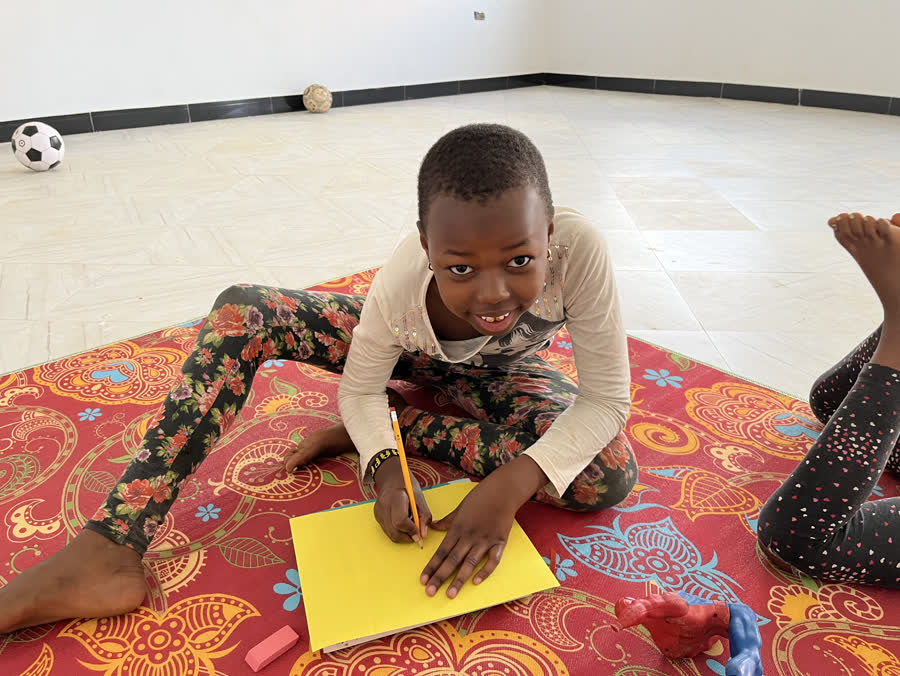 A girl lying on a red patterned mat, drawing on a yellow sheet of paper.