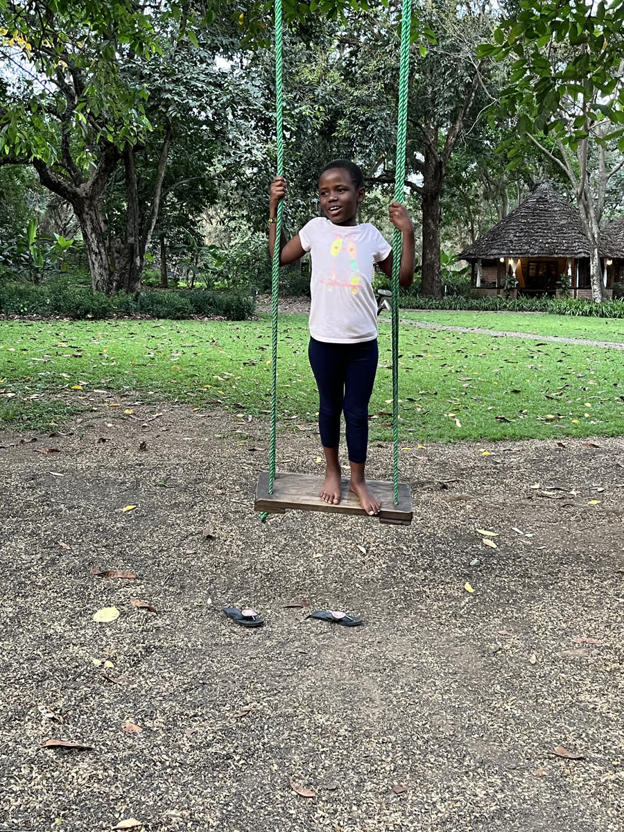 A girl standing barefoot on a rope swing under the trees, sandals left on the ground.