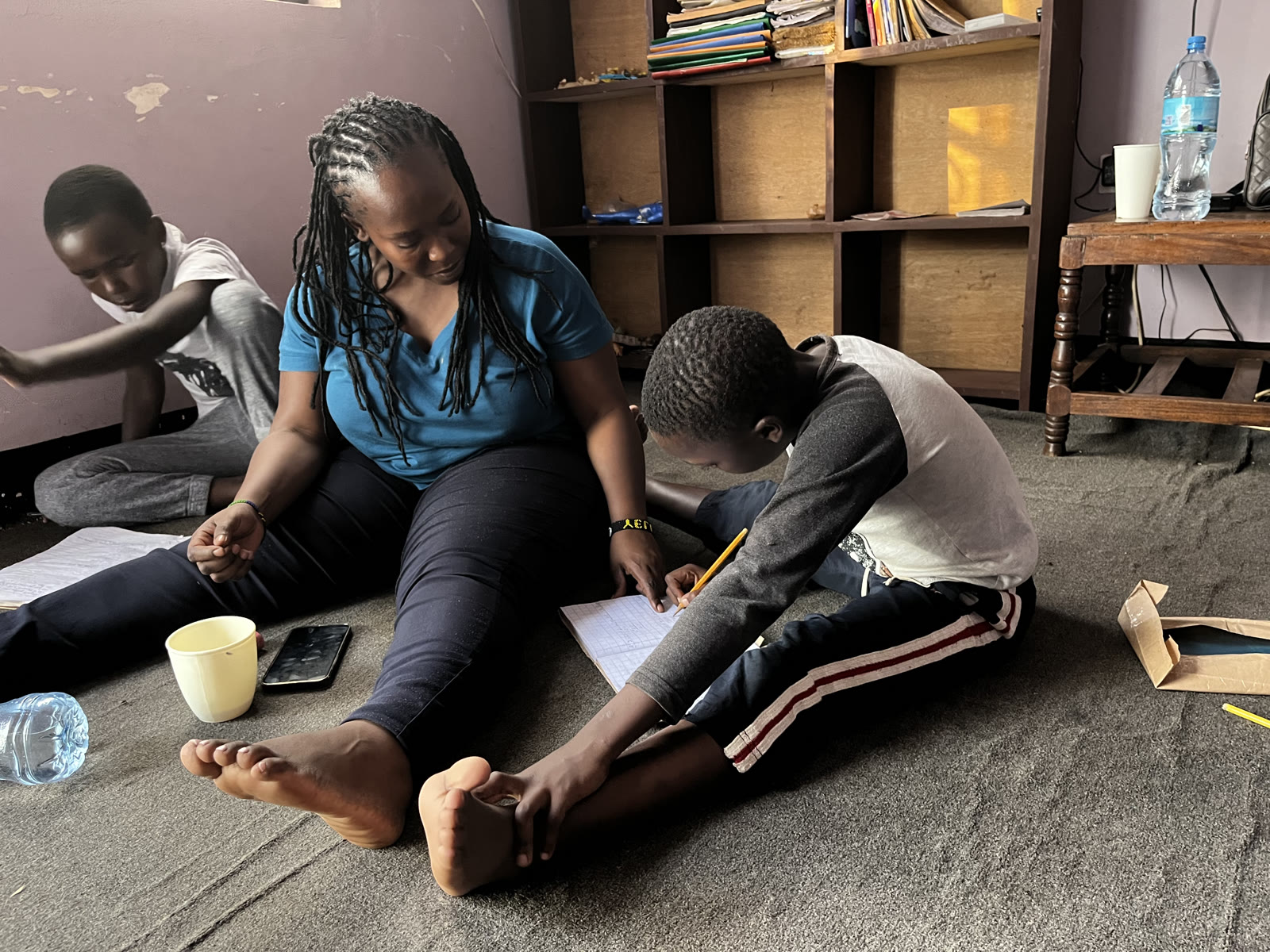Naomi Albert Ndesa sitting on the floor of the home helping a boy with his homework while another child works nearby.