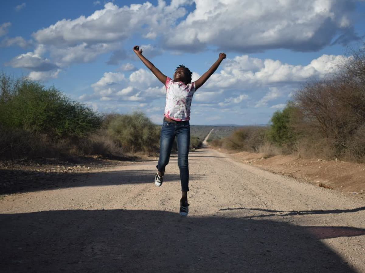 A young woman jumping with both arms raised on an open dirt road outside Arusha, under a wide sky.