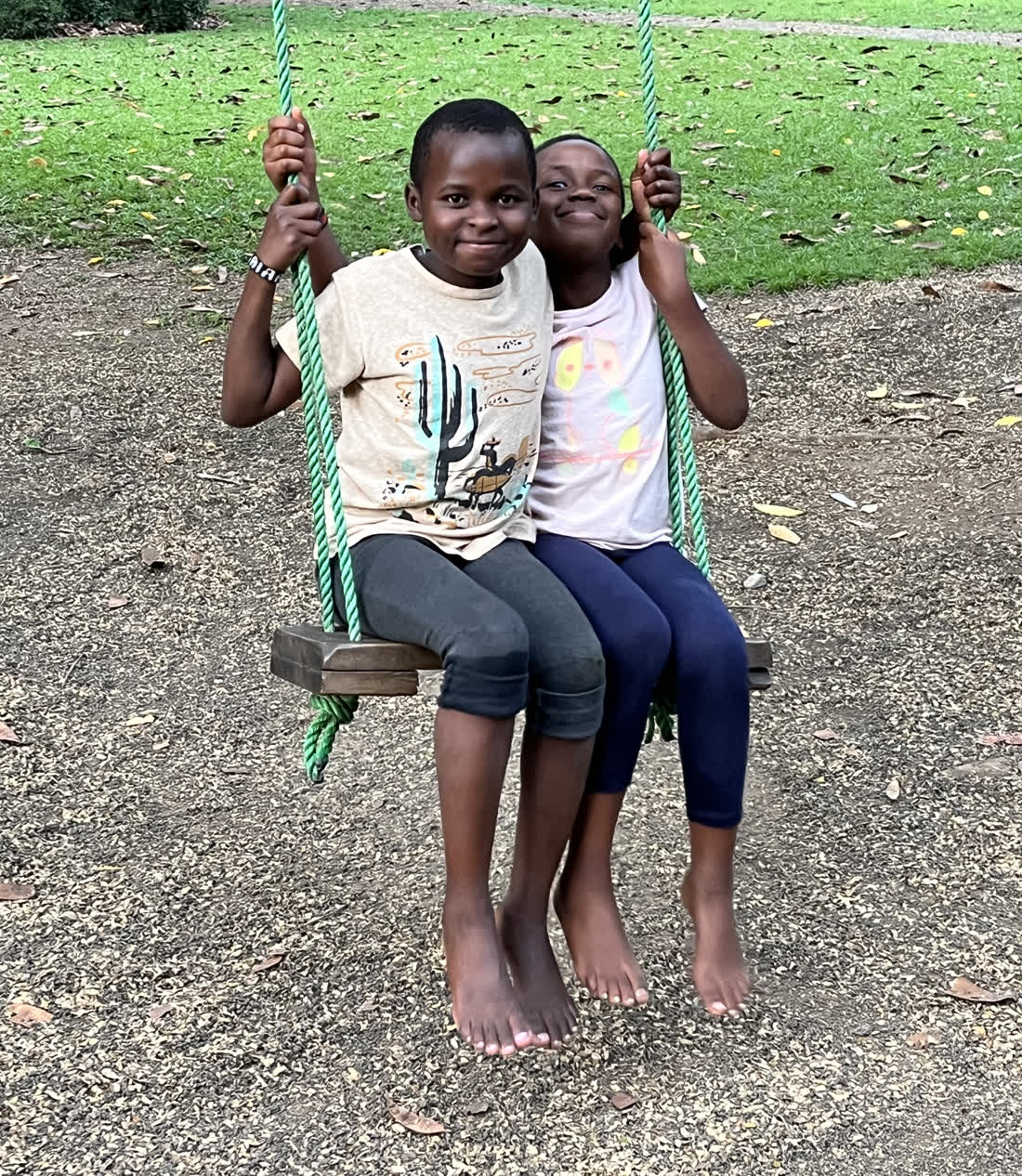 Two children from Holley Children Orphanage sharing one swing in the garden, holding the ropes and laughing.